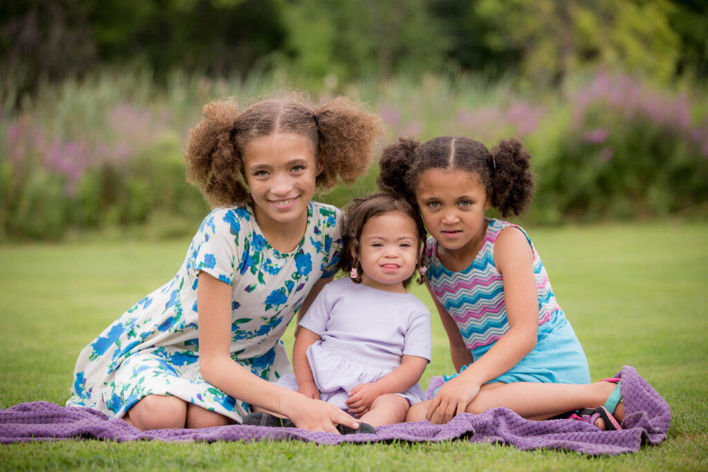 Three little girls sitting on a blanket in the grass.